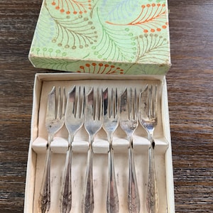 May include: A set of six vintage silver forks in a cream-colored box. The forks have a decorative pattern on the handles and are arranged neatly in the box. The box has a green and orange floral design on the lid.