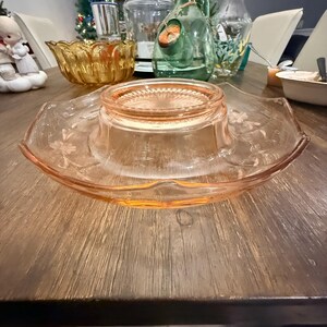 May include: A pale pink depression glass serving plate featuring a raised central pedestal base with a sunburst pattern and a wide, scalloped rim decorated with etched floral motifs. The glass has a translucent finish and a smooth, curved edge.