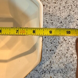 May include: A cream-colored, octagonal ceramic dish with a brown rim. The dish is approximately 9 inches long, as measured by a yellow tape measure. The dish is on a speckled gray surface.
