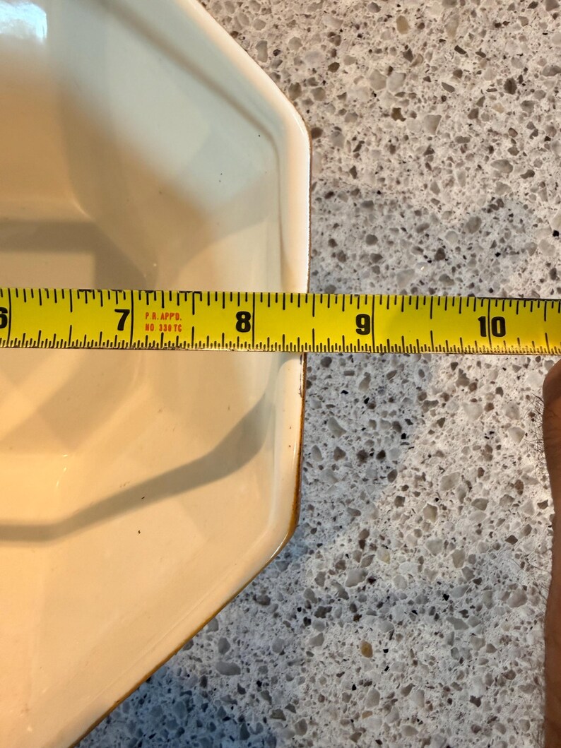 May include: A cream-colored, octagonal ceramic dish with a brown rim. The dish is approximately 9 inches long, as measured by a yellow tape measure. The dish is on a speckled gray surface.