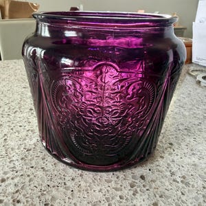 May include: A deep purple glass vessel featuring a wide, open top and a rounded body. The exterior surface is decorated with intricate, embossed scrollwork and floral patterns that wrap around the sides, creating a textured, reflective finish.