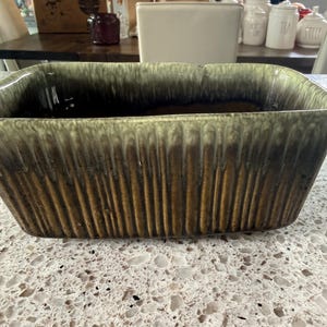 Pode incluir: Um vaso retangular de cerâmica com uma textura canelada vertical no exterior. O esmalte transita de um castanho escuro profundo na base para um verde sálvia claro e suave na borda superior, criando um gradiente de cor distinto e visível.