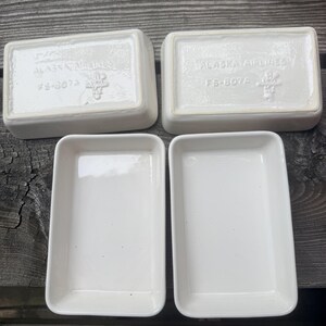 May include: Four rectangular white ceramic dishes. Two are positioned upside down, showing the embossed text Alaska Airlines, the codes FS-8073 and FS-807A, and a small logo. The other two dishes are placed right-side up, revealing their smooth interior.