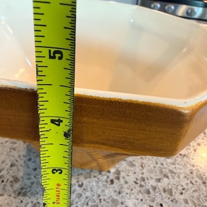 May include: A rectangular, off-white ceramic baking dish with a brown rim. The dish is approximately 5 inches deep. A yellow tape measure is positioned next to the dish, showing the depth.