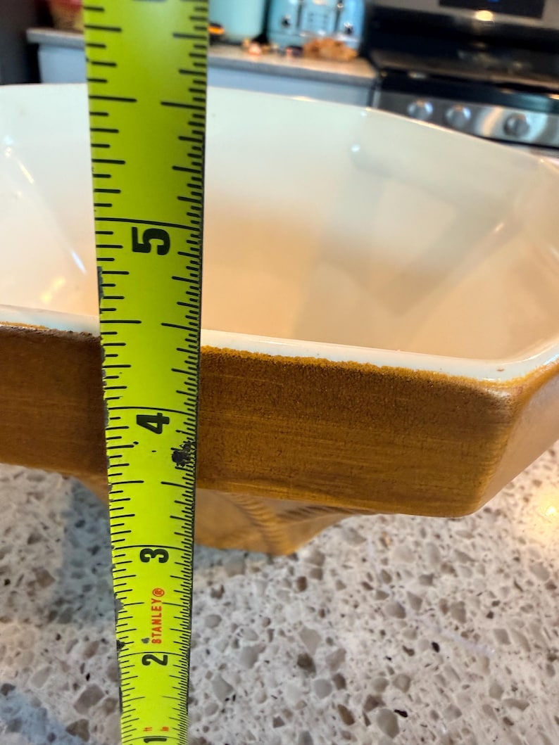 May include: A rectangular, off-white ceramic baking dish with a brown rim. The dish is approximately 5 inches deep. A yellow tape measure is positioned next to the dish, showing the depth.