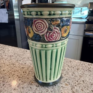 May include: A ceramic vase featuring a flared rim with a dark brown edge, a band of embossed pink roses and yellow flowers, and a lower section with vertical green fluted columns. The base is finished with a dark brown trim matching the rim.