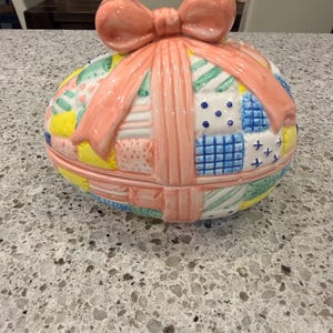 May include: A ceramic egg-shaped trinket box with a patchwork design in pastel colors. The box is adorned with a large pink bow on top and matching ribbon detailing. The lid and base are separate.