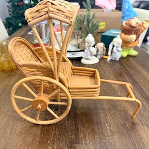 May include: A miniature, handcrafted rickshaw made of light brown woven wicker and wood. The decorative piece features a canopy, seat, and spoked wheels. The rickshaw is a charming, vintage-style home decor item.