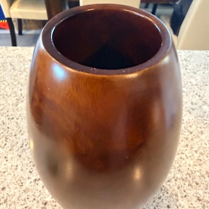 May include: A smooth, barrel-shaped wooden vase with a polished dark brown finish. The vase features a wide, circular opening at the top and a rounded, bulbous body that tapers slightly toward the base, showcasing a rich, natural wood grain pattern.