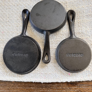May include: Three round black cast iron skillets arranged in a cluster. Two of the skillets feature the word Galerie embossed on the bottom surface, while the third skillet is positioned in the centre with its handle pointing downwards.