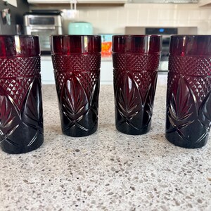 Op de afbeelding: Een set van vier dieprode glazen drinkbekers met een gestructureerd ruitpatroon op het bovenste gedeelte en een reeks verticale, bladachtige reliëfpatronen rond de basis, wat elk cilindrisch drinkglas een gefacetteerd uiterlijk geeft.