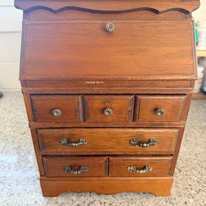 May include: A small, vintage wooden desk with a hinged top and multiple drawers. The desk is made of a warm-toned wood and features ornate metal hardware. The desk measures approximately 12 inches wide, 8 inches deep, and 14 inches tall.