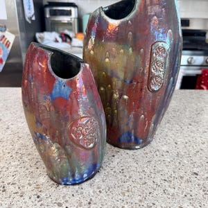 May include: Two ceramic vases with a unique, organic shape. The vases have a glossy, iridescent finish with colors of red, blue, and gold. The larger vase has a rectangular embossed design. The smaller vase has a circular embossed design.