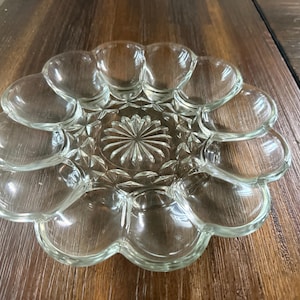 May include: A clear glass egg serving platter featuring a circular design with twelve individual oval-shaped indentations arranged around a central starburst pattern. The plate has a scalloped outer edge and a smooth, transparent finish.