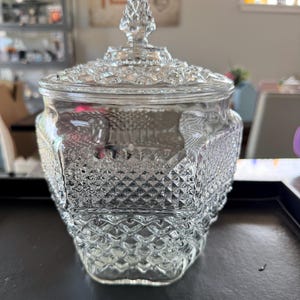 Peut inclure: Un bocal en verre transparent avec un couvercle assorti, présentant un motif en losanges texturé sur tout le corps et le couvercle. Le couvercle est surmonté d'un fleuron pointu et facetté. Le bocal a une forme large et arrondie.