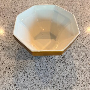 May include: An octagonal ceramic bowl with a cream-colored interior and a golden-yellow rim. The bowl's geometric design and neutral color palette make it a versatile piece for serving or display. The bowl is empty.