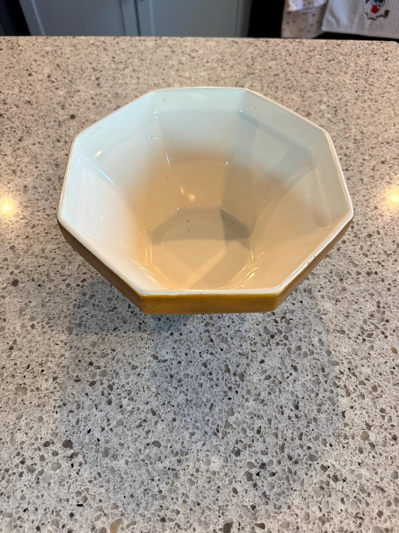 May include: An octagonal ceramic bowl with a cream-colored interior and a golden-yellow rim. The bowl's geometric design and neutral color palette make it a versatile piece for serving or display. The bowl is empty.