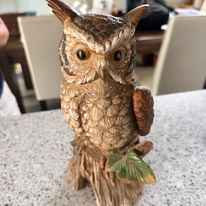 May include: A ceramic owl figurine perched on a textured tree stump base. The owl features brown and tan feathers, large amber-colored eyes, and pointed ear tufts. A single green leaf is visible on the side of the stump base below the owl.