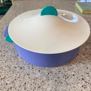 May include: A round, two-toned serving dish with a white lid and a lavender base. The lid has a teal handle and teal side tabs. The dish is designed for serving food.
