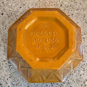 May include: An octagonal, amber-colored ashtray with a textured rope design on the sides. The base is a solid amber color with the text "HAEGER NO. 834 U.S.A." embossed. A vintage decorative piece.