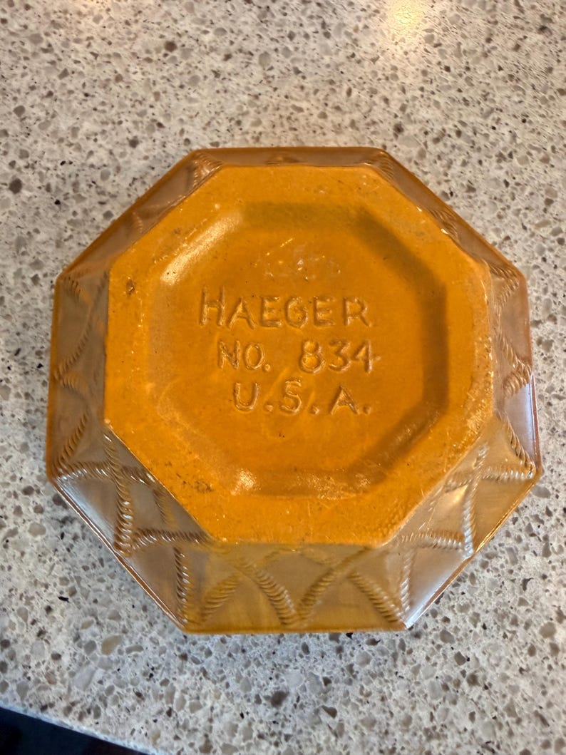 May include: An octagonal, amber-colored ashtray with a textured rope design on the sides. The base is a solid amber color with the text "HAEGER NO. 834 U.S.A." embossed. A vintage decorative piece.