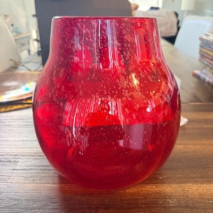 May include: A round, red glass vase with a wide base and a narrower neck. The vase has a speckled texture and a glossy finish. The red colour is vibrant. This decorative vase is suitable for displaying flowers or as a standalone art piece.