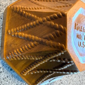 May include: A brown, hexagonal ceramic container with a rope-like design. The container has a glossy finish and a label that reads "Haeger" and "U.S.A.". The rope design is raised and creates a textured surface.