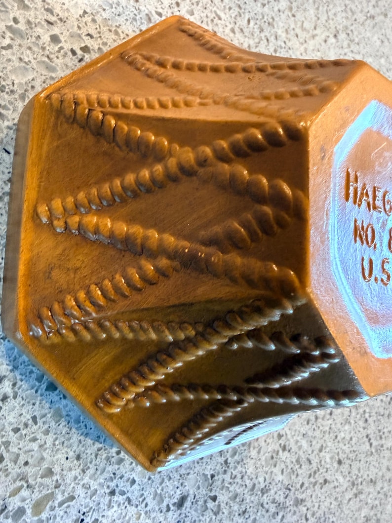 May include: A brown, hexagonal ceramic container with a rope-like design. The container has a glossy finish and a label that reads "Haeger" and "U.S.A.". The rope design is raised and creates a textured surface.