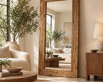 Puede incluir: Un espejo de pie alto y rectangular con un marco grueso de madera marrón desgastada con una textura de veta visible. El espejo está colocado verticalmente contra una pared, reflejando un espacio de sala con muebles y decoración.