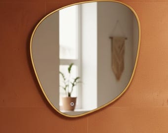 May include: An irregularly shaped mirror with a gold-colored frame. The mirror is mounted on a textured, reddish-brown wall. The reflection shows a window with a potted plant and a decorative wall hanging.