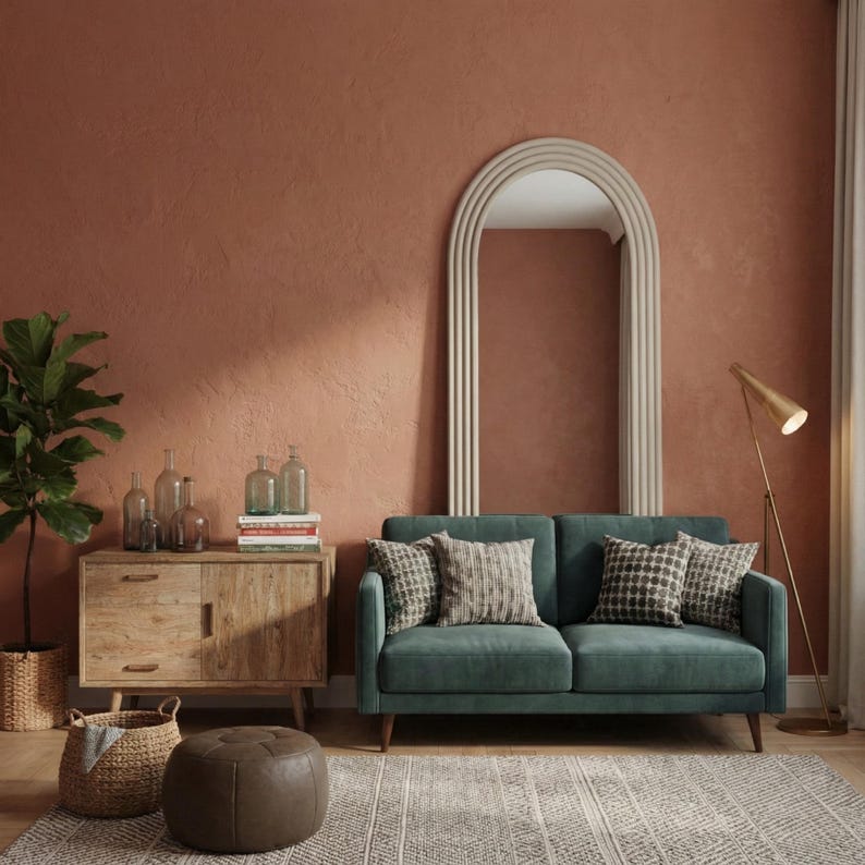 May include: A teal two-seater sofa with patterned throw pillows sits in front of a large arched mirror. A wooden cabinet with glass bottles and a floor lamp are also in the room. The wall is painted a warm terracotta color.
