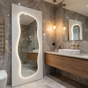 Puede incluir: Un baño moderno con un espejo grande, blanco, de forma orgánica con iluminación integrada. La habitación cuenta con un mueble de baño de madera con un lavabo rectangular blanco, accesorios dorados y baldosas grises con efecto mármol.