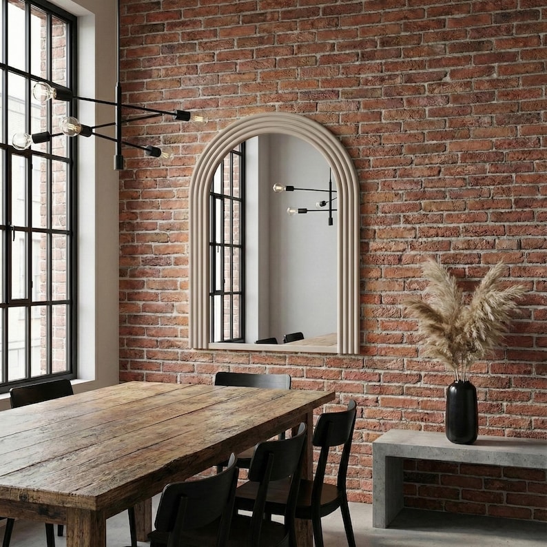May include: A dining room with a large wooden table and black chairs. A beige arched mirror hangs on a red brick wall. A black light fixture and a vase of dried plants complete the decor.