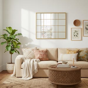 May include: A living room setting with a beige sofa, cushions, and a woven throw. A round wooden coffee table is in front of the sofa. A large gold-framed mirror hangs on the wall above the sofa, and a potted plant is to the left.