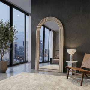 May include: A large, arched floor mirror with a light wood frame stands in a modern room. The mirror reflects a view of a city skyline. A small marble side table with a lamp and a leather chair are nearby.