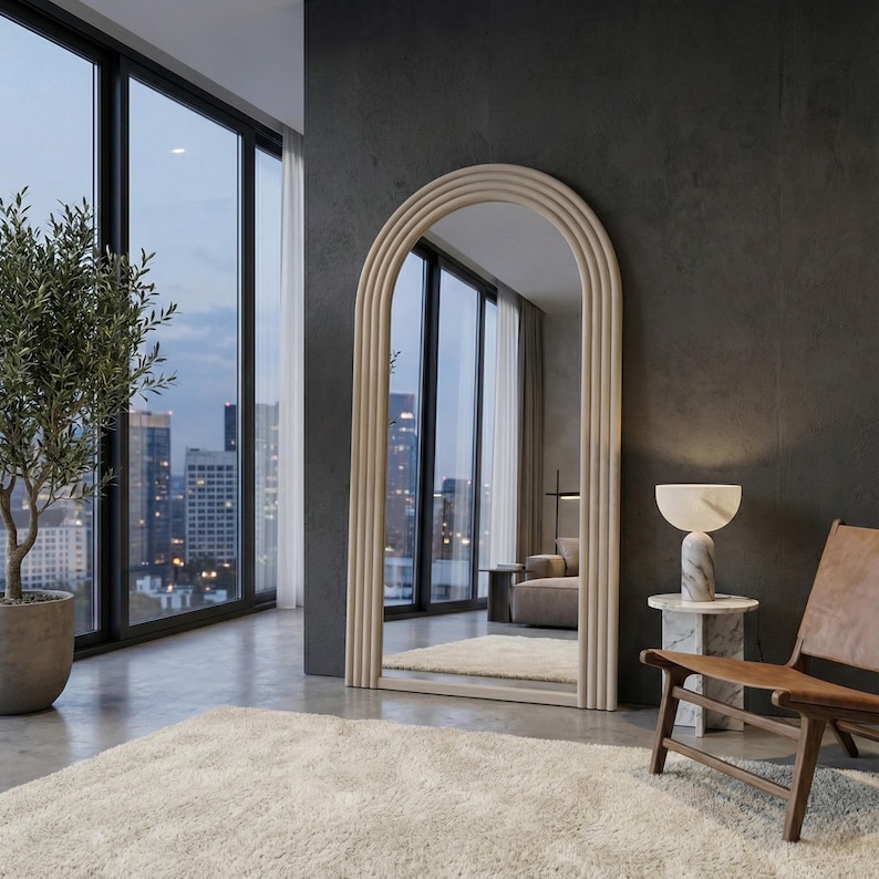 May include: A large, arched floor mirror with a light wood frame stands in a modern room. The mirror reflects a view of a city skyline. A small marble side table with a lamp and a leather chair are nearby.