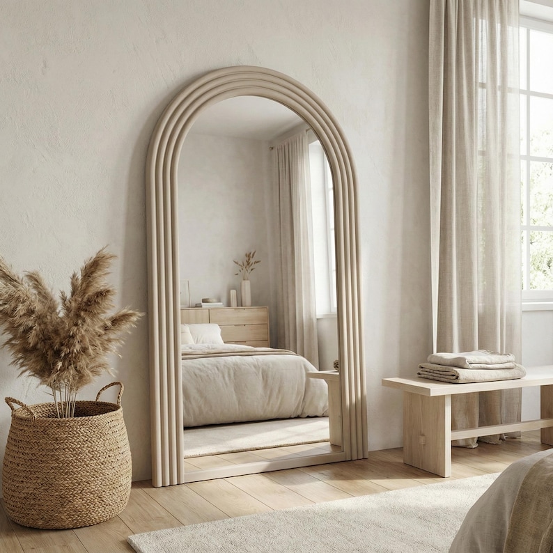 May include: A large, arched, full-length mirror with a light wood frame. The mirror reflects a bedroom with neutral tones, a bed, and a woven basket with dried plants. The frame has a layered, vertical design, adding a decorative element.