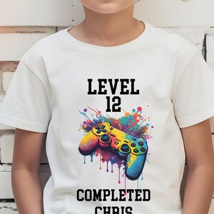 May include: White t-shirt featuring a colorful graphic of a video game controller with the text "LEVEL 12 COMPLETED CHRIS". The controller has yellow, blue, and green accents, with a splash of colorful paint behind it. The shirt is likely for a child.
