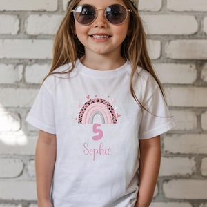 May include: A white short-sleeved t-shirt featuring a graphic of a rainbow with pink, grey, and leopard print arches, surrounded by small pink and black hearts. Below the rainbow, the number 5 and the name Sophie are printed in pink script.