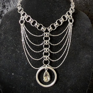 Puede incluir: Collar plateado con un diseño en capas. El collar presenta múltiples hilos de cadena que caen en cascada desde la parte superior, conectados por eslabones circulares. Un gran colgante de anillo sostiene un cristal facetado en forma de lágrima. Se muestra en un maniquí negro.