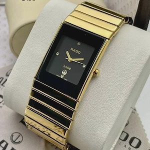 Peut inclure: Une montre-bracelet Rado Jubilé avec un cadran rectangulaire noir, des aiguilles dorées, quatre index en diamant et l'inscription Rado et jubilé. La montre possède un bracelet à maillons en métal doré poli avec des accents noirs.