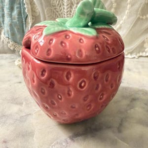 May include: A ceramic strawberry-shaped jar with a removable lid. The jar features a glossy red finish with textured indentations representing seeds and a sculpted green leaf handle on top. The lid fits securely onto the base of the container.