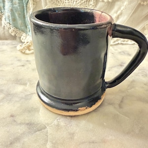 May include: A glossy black ceramic mug with a sturdy handle and a wide base. The mug features a smooth, dark finish that covers the exterior and interior, while the bottom edge reveals a natural, unglazed tan clay rim with a small chip.