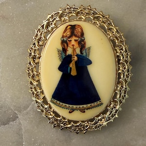 May include: An oval brooch featuring a gold-toned filigree frame surrounding a cream-colored center. The center displays an illustration of an angel with brown hair, wearing a long dark blue dress and playing a gold-colored trumpet instrument.