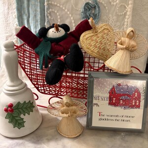 May include: A red wire sleigh containing a fabric snowman, a woven straw heart, and a straw angel. Beside it sits a white ceramic bell with holly, a second straw angel, and a framed print of a red house with the text, The warmth of Home gladdens the Heart.