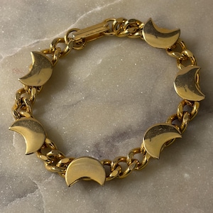 May include: A gold-toned chain link bracelet featuring five evenly spaced, smooth crescent moon charms. The bracelet is constructed with thick, interlocking oval links and includes a secure rectangular clasp mechanism at the top.