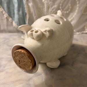 May include: A cream-colored ceramic pig-shaped vessel with a speckled finish, featuring a cork stopper in the snout, small ears, and a series of circular holes along the top of the back. The object is positioned horizontally on its side.