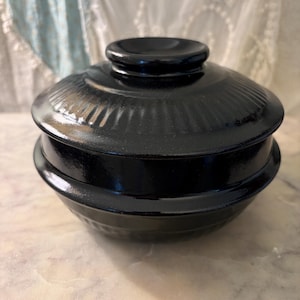 May include: A glossy black lidded bowl featuring a smooth, rounded silhouette with a raised circular handle on top. The lid and the base of the bowl are decorated with a subtle, repeating vertical ribbed pattern that encircles the entire exterior.