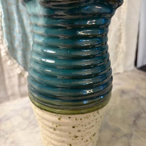 May include: A ceramic vase featuring a two-tone design with a glossy, deep teal upper section and a matte, off-white speckled lower section. The body of the vase has a horizontal ribbed texture throughout, and the rim has a wavy, flared shape.