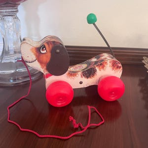 May include: Vintage pull-along toy dog with a brown, white, and pink body, black ears, and red wheels. The dog has a red pull string and a green ball on a spring. A classic children's toy.